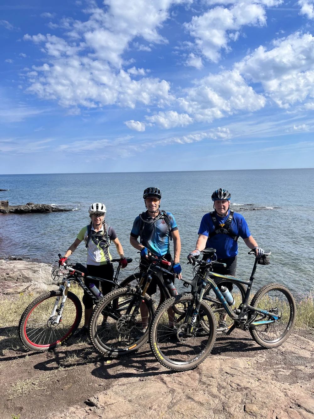 Three helmeted cyclists with their bicycles beside a rocky shoreline